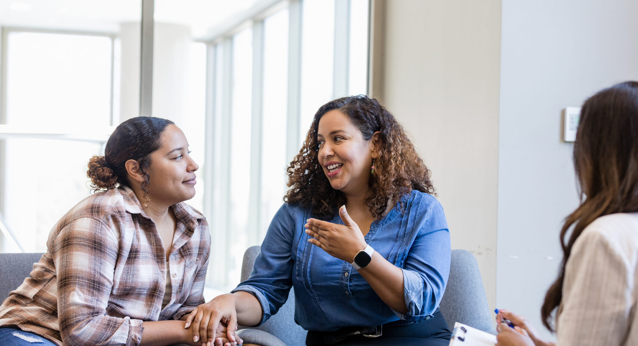 Women’s Health Spotlight: Pelvic Health After Childbirth Two women in casual clothing hold hands during a counseling session. A therapist takes notes.