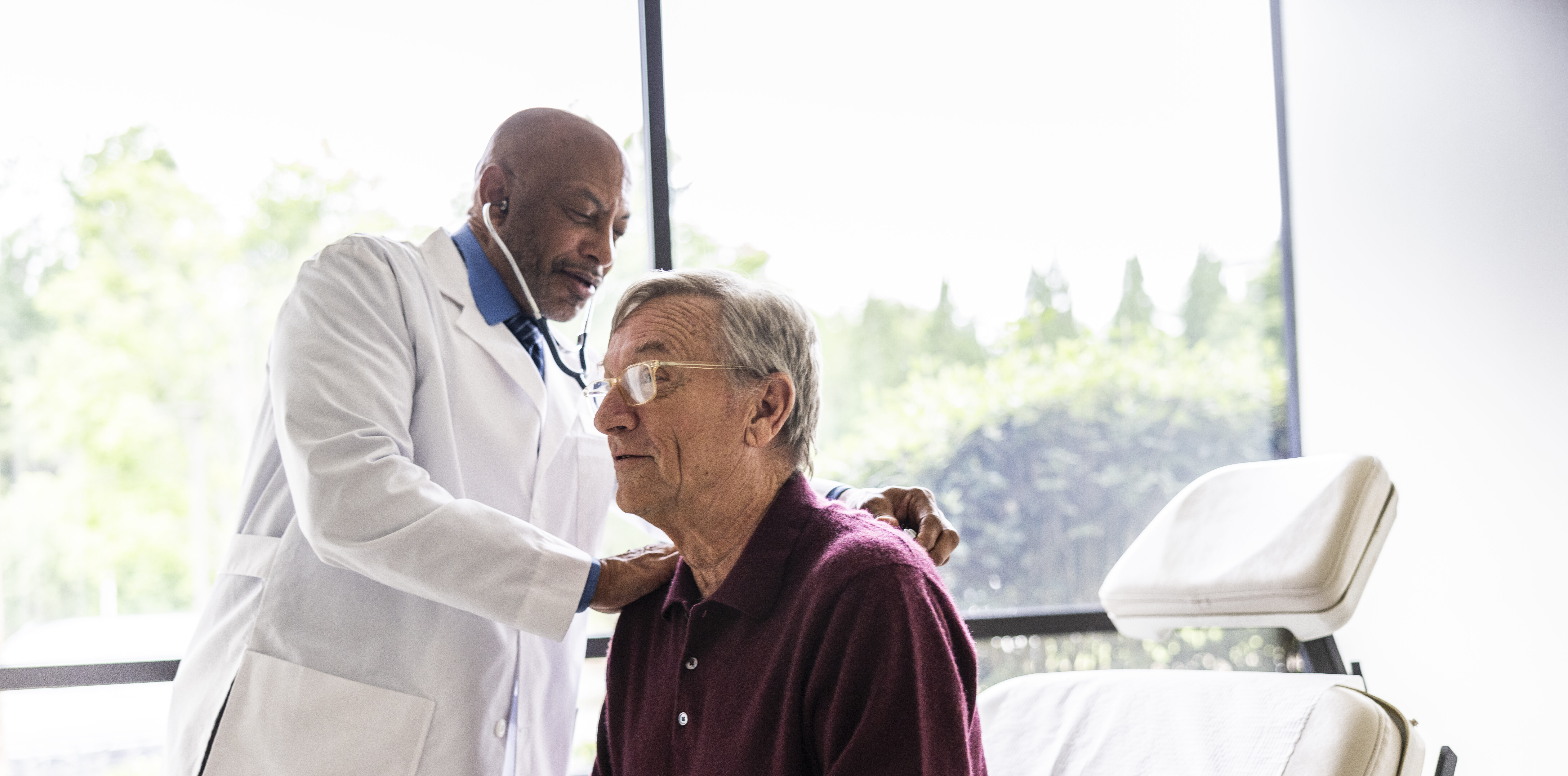 Male doctor listening to senior man's heart in exam room