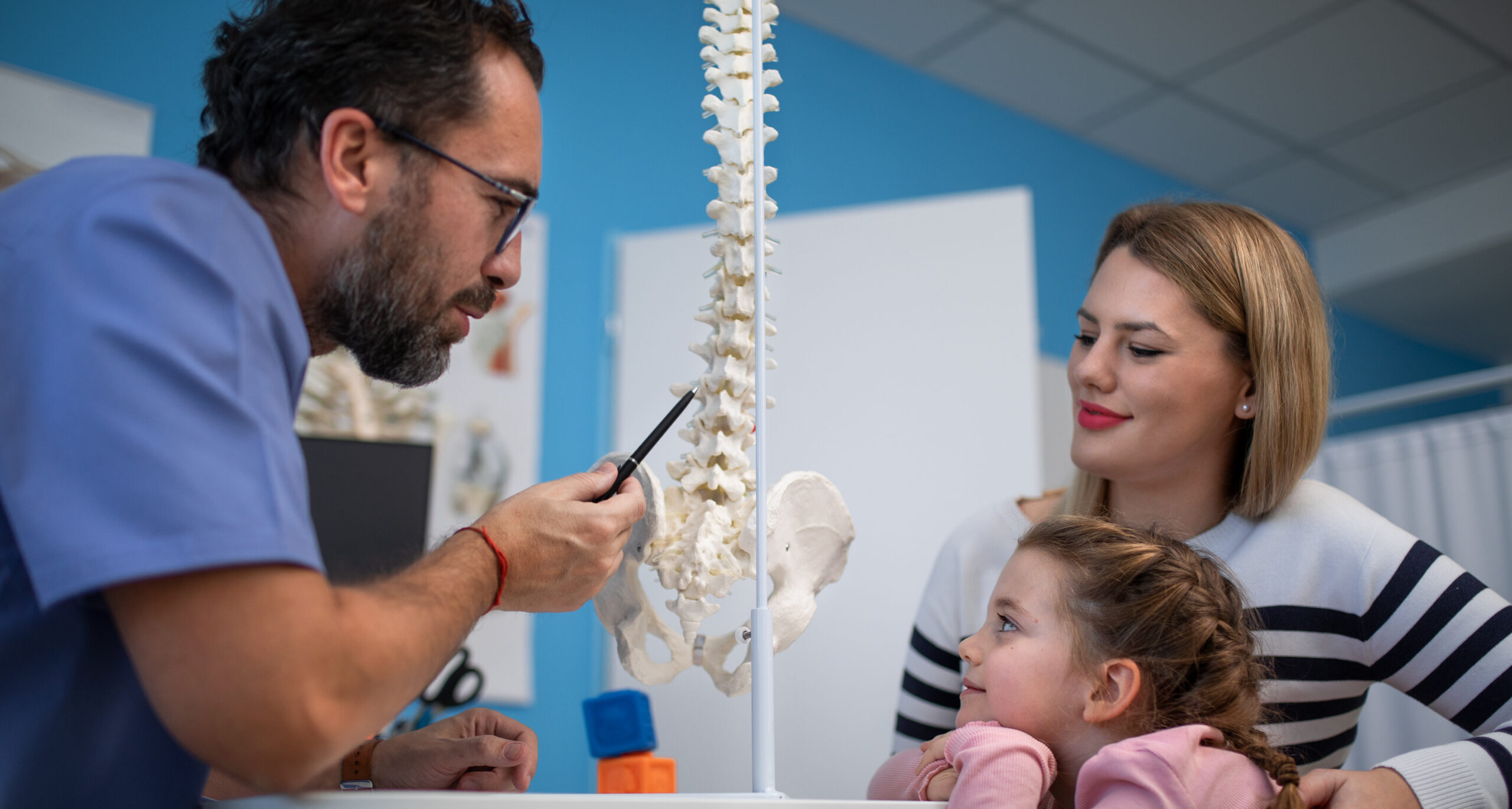 Orthopedist doctor showing skeleton model to his little patient with her mother in his office.