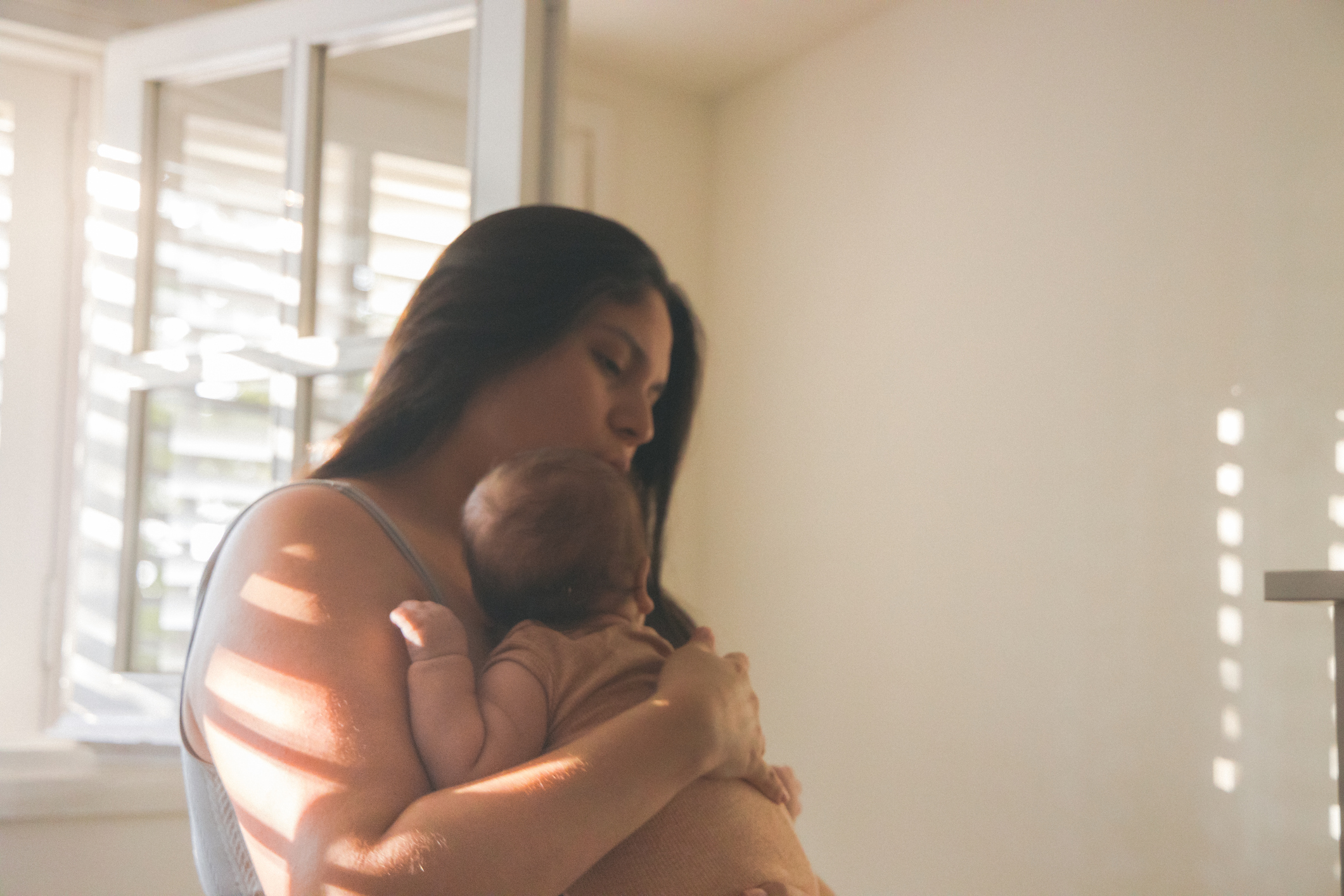 Mother cuddling newborn baby at home in warm morning sunlight after a sleepless night